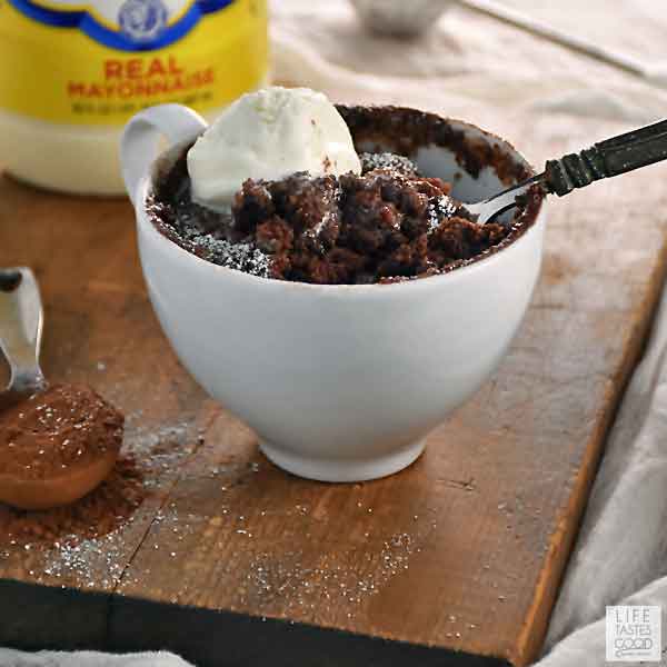 White mug filled with gooey chocolate mug cake set on a rustic wooden board. A spoon is scooping out some gooey chocolate cake.