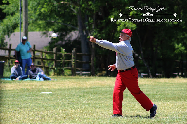 Man  in a grassy field dressed in an 1800s baseball uniform. He is wearing red slacks, a gray button down dress shirt with long sleeves and has some letter on the front in red. He is wearing a baseball cap in red and blue bill. The article is Vintage baseball by rosevinecottagegirls.com