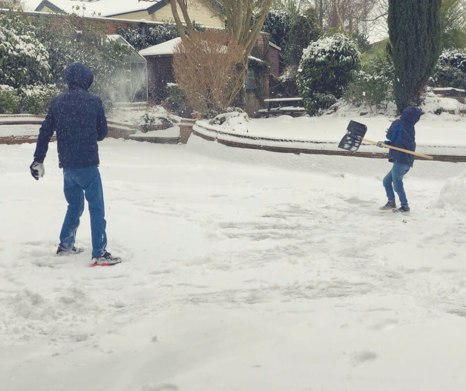 A teen boy and a tween boy play in the snow. The ground is covered in 2 inches of snow, footprints and drag lines can be seen in the snow. In the background are snow-covered trees and leaves. The older boy is throwing a snowball at the younger boy while the younger boy is holding a snow shovel to protect himself.  Image for "Post Comment Love 2nd - 4th March"