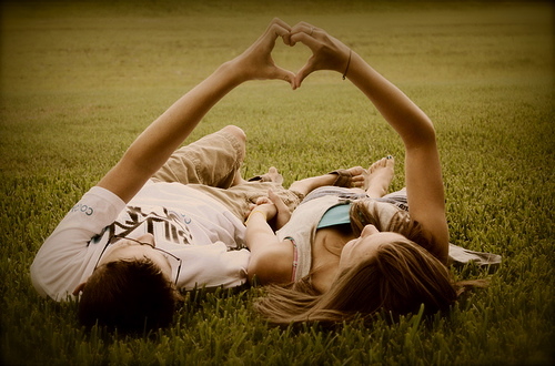 Pareja de chico y chica tumbados en el campo, haciendo un corazón con las manos.