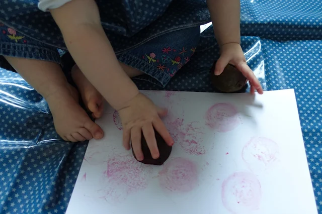 A baby making stamps with cut beetroot on a white piece of card on a wipe clean messy mat in blue with small white stars