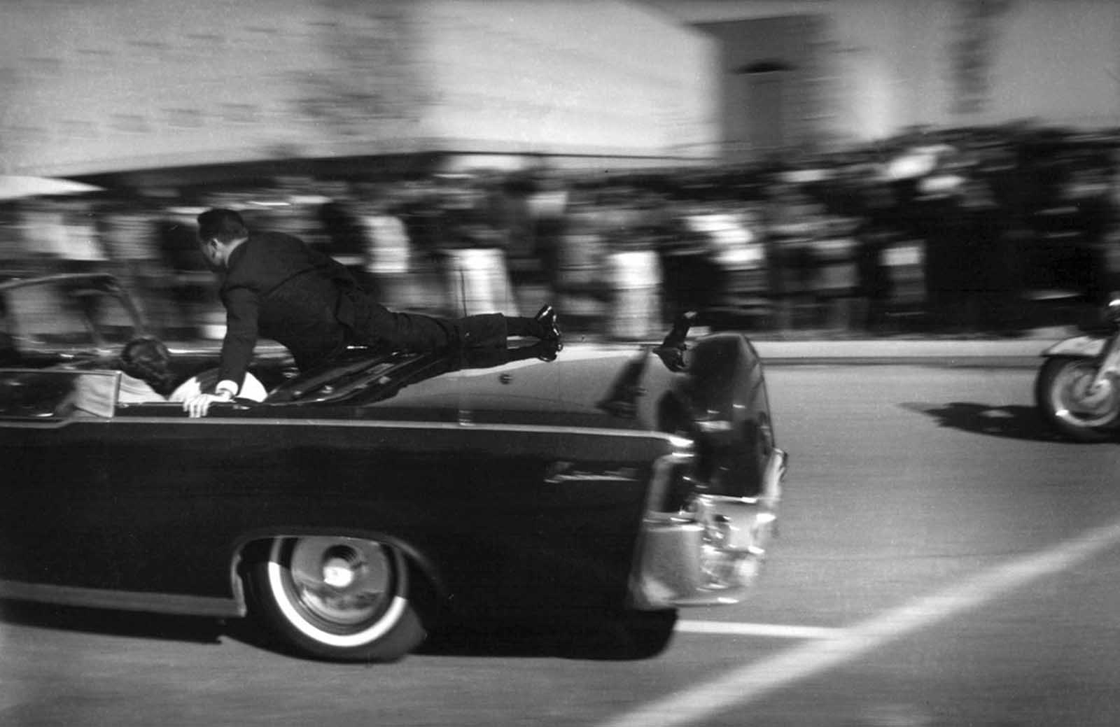 Seconds after shots rang out, the limousine carrying the mortally wounded President John F. Kennedy races toward the hospital in Dallas, Texas, on November 22, 1963. Secret Service agent Clinton Hill is riding on the back of the car, Nellie Connally, wife of Texas Gov. John Connally, bends over her wounded husband, and first lady Jacqueline Kennedy leans over the president. 