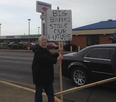 Older woman holding a sign along a highway across from a Bank of America branch. It reads Big Banks Stole Our Future