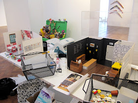 Two dolls' house miniature scenes, being built on  a table in a gallery during install.
