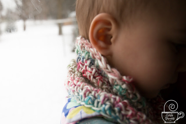 Image from the side of a multi-colored cowl on a toddler's next. Toddler's ear is visible. Behind the toddler is a snowy winter wonderland.
