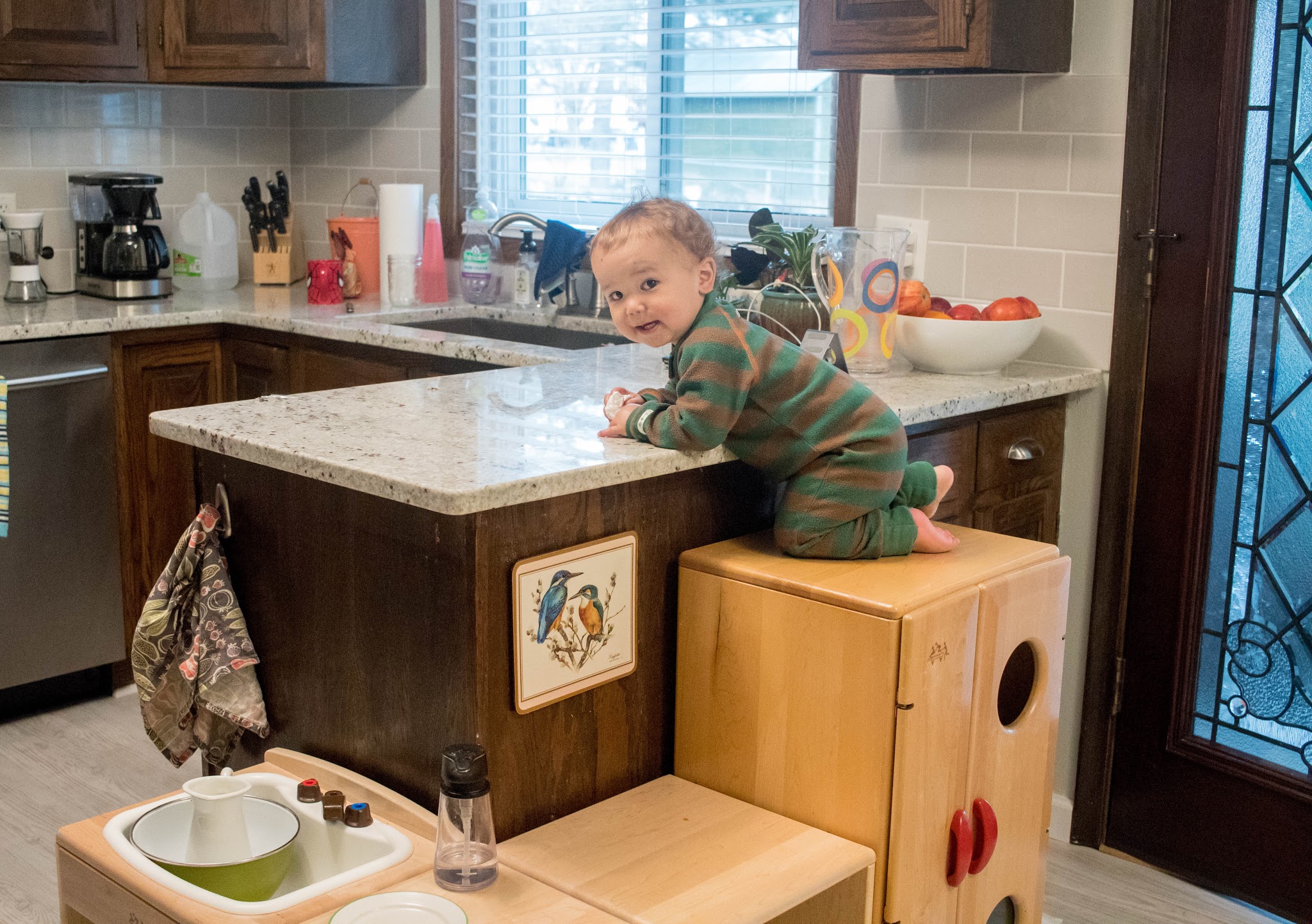 A young toddler climbs on kitchen counters of Montessori home. He looks toward the camera as his mom says "feet stay on the floor" as an example of positive action language to use when children test boundaries.