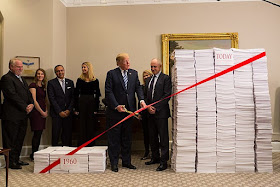 Photo shows President Trump, surrounded by aides and holding a pair of scissors, about to cut red ribbon in front of two piles of papers: a small pile labeled "1960" and a large pile labeled "today."