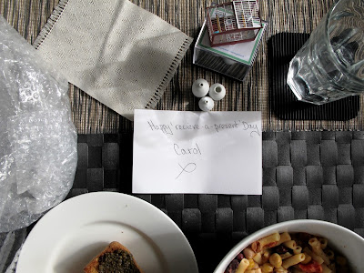 Dining table with a bowl of soup, a plate of pesto toast, a glass of water, bubble wrap and a selection of modern dolls' house miniatures next to a card on which is written 'Happy receive-a-present Day.'