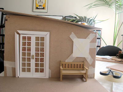 Side view of a taped-together modern dolls' house miniature kit, with a set of white french doors propped against the wall and a park bench next to them.
