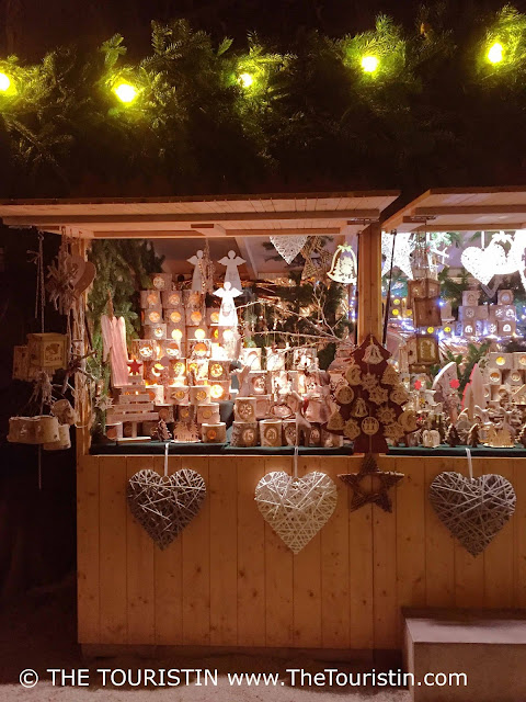 Three large-sized white wooden heart-shaped ornaments and a wooden woven star hang on the front of a romantically illuminated wooden stall selling Christmas ornaments, angels, and tea lights.