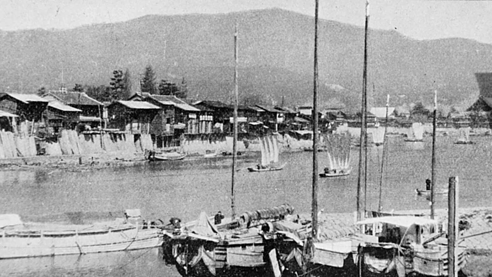 Looking north from the vicinity of the Aioi Bridge (the central T-shaped bridge targeted by the bomb). Wooden houses line the bank of the Otagawa, with traditional Japanese river boats in the foreground.