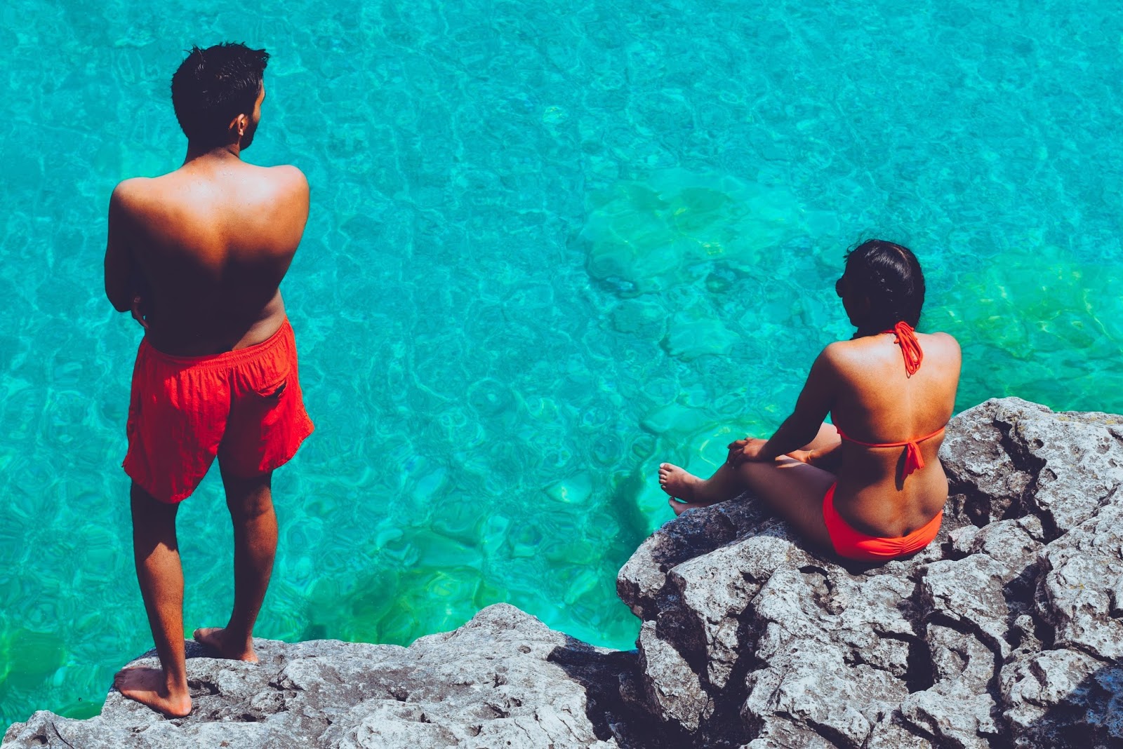 Man and a woman in red swimwear perched on a rock overlooking a bright blue sea