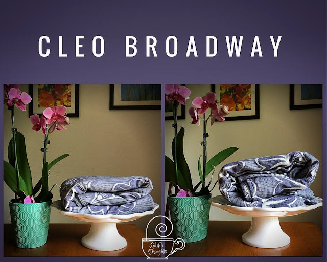 [Image is a graphic of two photos against a grayish-purple background. The text in white says the name of woven wrap featured, Cleo Broadway. Both images have a grayish-purple geofloral patterened woven wrap baby carrier neatly folded on top of a white ceramic cake plate next to a potted orchid plant, on a wood table. The left photo is of the woven wrap in "loom state." The right photo is of the woven wrap post-wash, also referred to as "bloom state" and it's folded the same way as the first photo but has poofed so it looks bigger and fluffier.]
