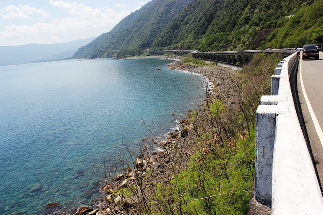 A view of the highway adjacent to the rocky shoreline and a view of the clear sea waters.