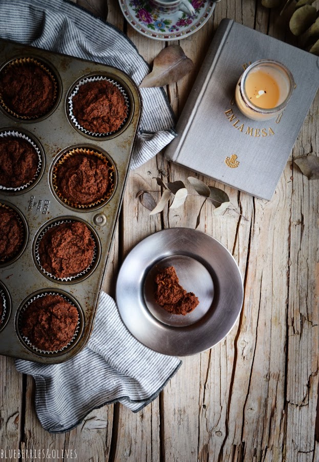 Plan cenital, bandeja de muffins horneados, msesa madera envejecida, paño de lino, plato con pedazo de muffin mordido,ramas eucalipto secas, plato y taza de café porcelana