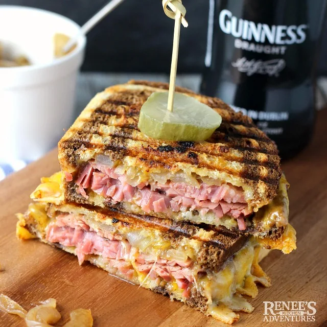 Corned Beef Panini with Caramelized Guinness Onions on a board, cut in half, with a pickle garnish