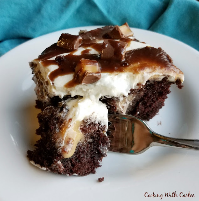 This chocolate cake is full of creamy, caramelly goodness, topped with fluffy frosting, drizzled with fudge and sprinkled with candy. Mmmmmm, it's good! piece of rolo poke cake on plate with chocolate cake, caramel filling, fluffy frosting, chocolate drizzle and bits of rolos
