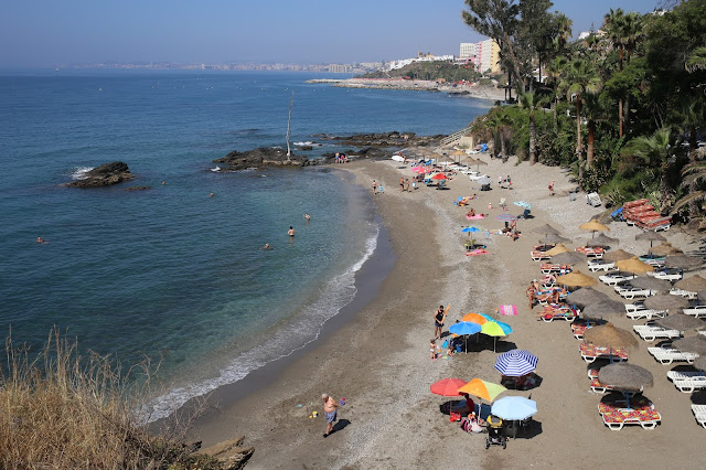Playa pequeña con gente sobre la arena, vegetación a su espalda, y las azules aguas del mar a su frente.