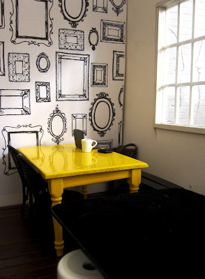 Corner of a modern one-twelfth scale miniature cafe with a yellow table under a window. Ont he table is a laptop, mug of coffee and a wallet.