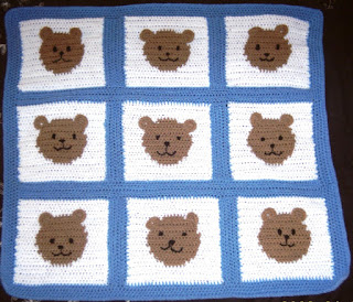 A square baby blanket made up of 9 squares, joined by blue borders. Each square is white with a tan circle in the middle. The circle is a teddy bear's face. Facial features are embroidered in black yarn. The ears are shaped like half circles and stitched on to the surface of the blanket along the top edge of the tan circles so that they can flap about.  The blanket resembles a tic tac toe game except there are faces where the noughts and crosses should be and the entire blanket is finished off with a plain blue border of solid crochet around the perimeter.