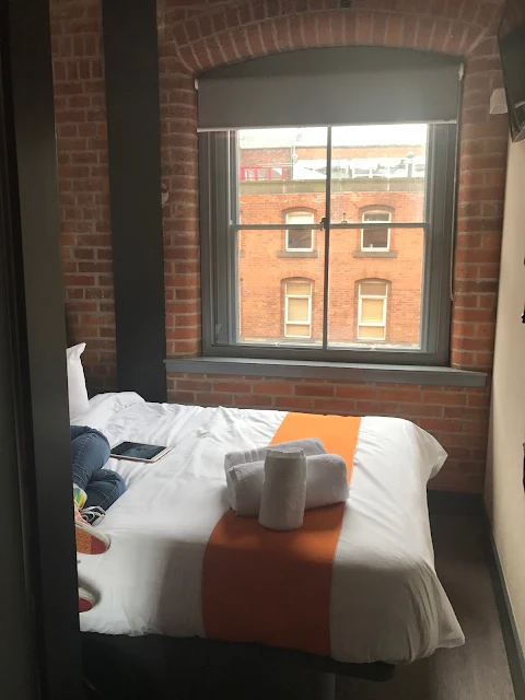 A double bed with an orange strip and a large window and not a lot of space