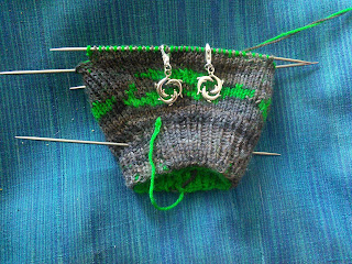 A double-knit mitten on double-pointed needles, knit in contrasting colours of green and brown yarn. There are two dolphin charm stitch markers on the needle, marking the increases for the thumb gusset. 