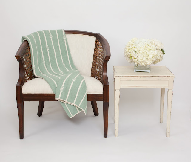 Nbaynadamas throw elegantly placed on on upholstered wood chair next to a white vintage side table holding a glass vase of white hydrangeas