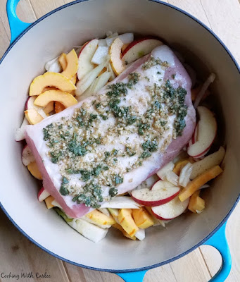 turquoise enameled dutch oven layered with sliced apples and vegetables with a hunk of herb coated pork loin on top ready to go in oven.