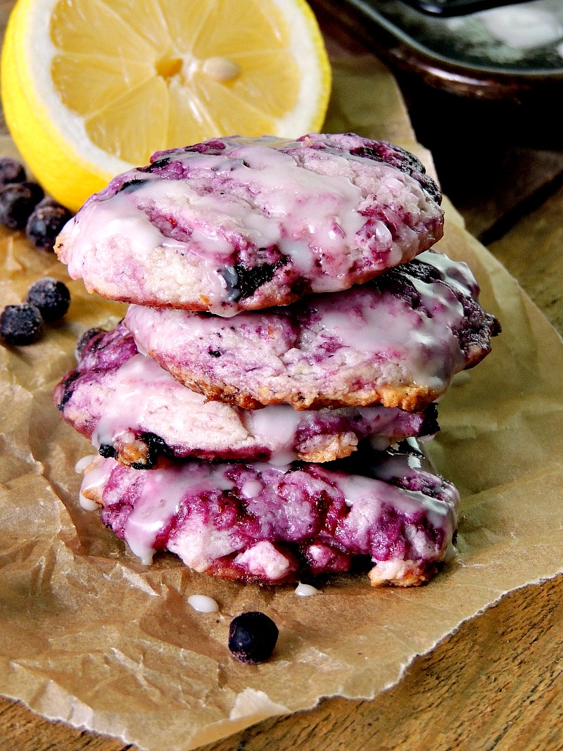 Blueberry Cheesecake Cookies with Lemon Glaze stacked on a piece of parchment paper with a lemon and blueberries scattered around.