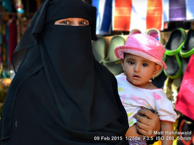 Street double portrait of a Muslim Indian woman with beautiful eyes, wearing a black niqab and burqa, and carrying her little daughter on her arms; Benaulim, South Goa, India. people; street portrait; headshot; face; India; Goa; Muslim lady; abaya; tourist; niqab; eye contact; eyes; Nikon DSLR D3100; Nikkor AF-S 50mm f/1.8G; portrait; double portrait; duo portrait