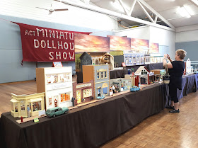 A woman photographing one of the one-twelfth scale buildings on display at a miniatures show. Behind the display hangs a fabric wallhanging that spells out 'ACT Miniature and Dollshouse show'.