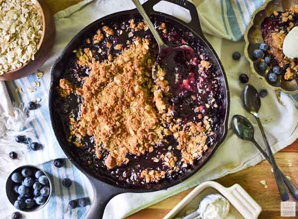 Blueberry Crisp in a cast iron skillet with fresh blueberries on the side and 2 spoons