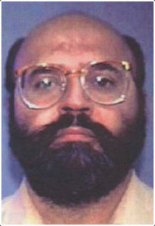 Interpol mug shot of a balding, middle-aged man of Indian ethnicity. He has a fringe of black hair and a full black beard and mustache and is wearing oversized 1980s style glasses.
