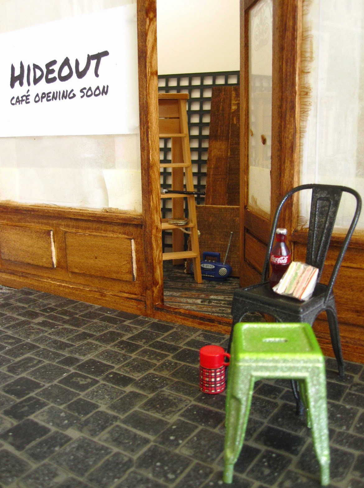 Modern dolls' house miniature cafe under renovation, with builder's lunch on a stool outside the front door.