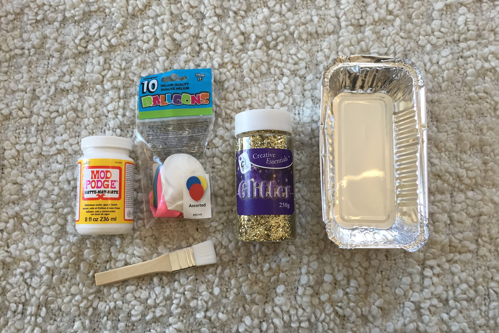 a selection of supplies for a DIY glitter bowl, including matte mod podge, multicoloured balloons, a wide brush, a tub of gold glitter, and a silver foil takeaway tray