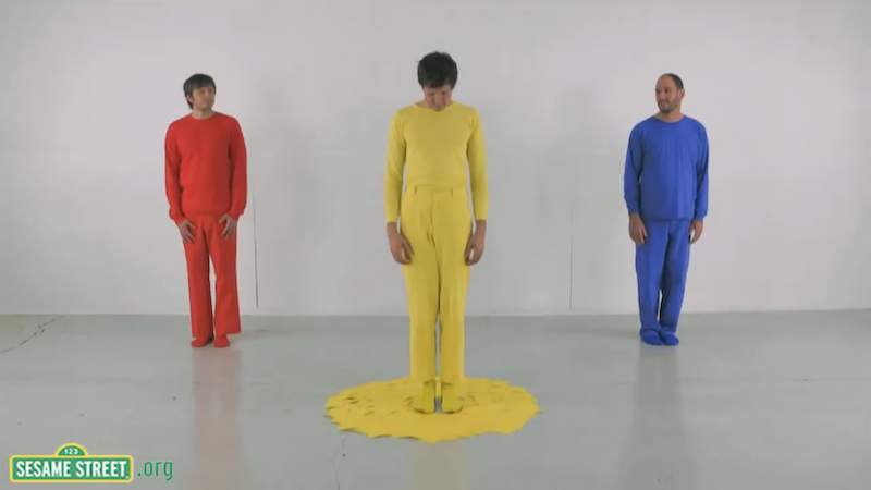 Three members of OK Go dressed in bright red, yellow, and blue respectively, the yellow one with a puddle of yellow paint at his feet