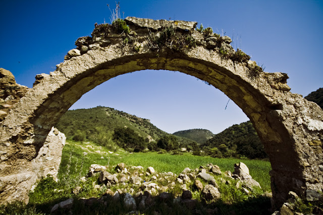 Un arco de pieda en ruinas, probablemente medieval, a través del cual se puede ver en la  sierra andaluza verde, crecida de hierba por doquier.