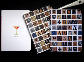 Three notebooks: one is white with a martini glass drawn on the cover. Two are covered with fabric printed with various letter As in wood tones.