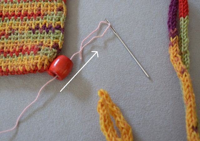 Close-up of bead on the bottom right corner of bag. A wool needle has been threaded and pushed through the bead from the bottom towards the top, leaving the tail of the yarn remaining inside the bead.