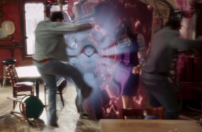 Janet stands in front of the makeshift demon portal (metal frame with watery red electricity in the middle) and has punched away two demons with her afterlife powers. There's a radiant light blue circle that indicates the supernatural strength with which she has punched the two demons, who are both flying backwards.