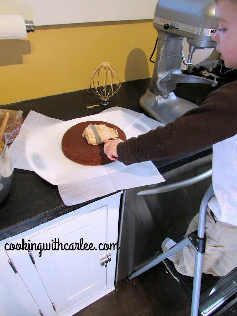 little dude standing on a stool and spreading frosting over the first layer of cake