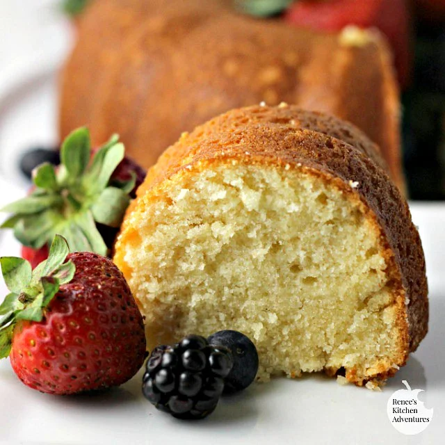 Slice of Buttermilk Pound Cake on white plate with berries in foreground, whole cake in background