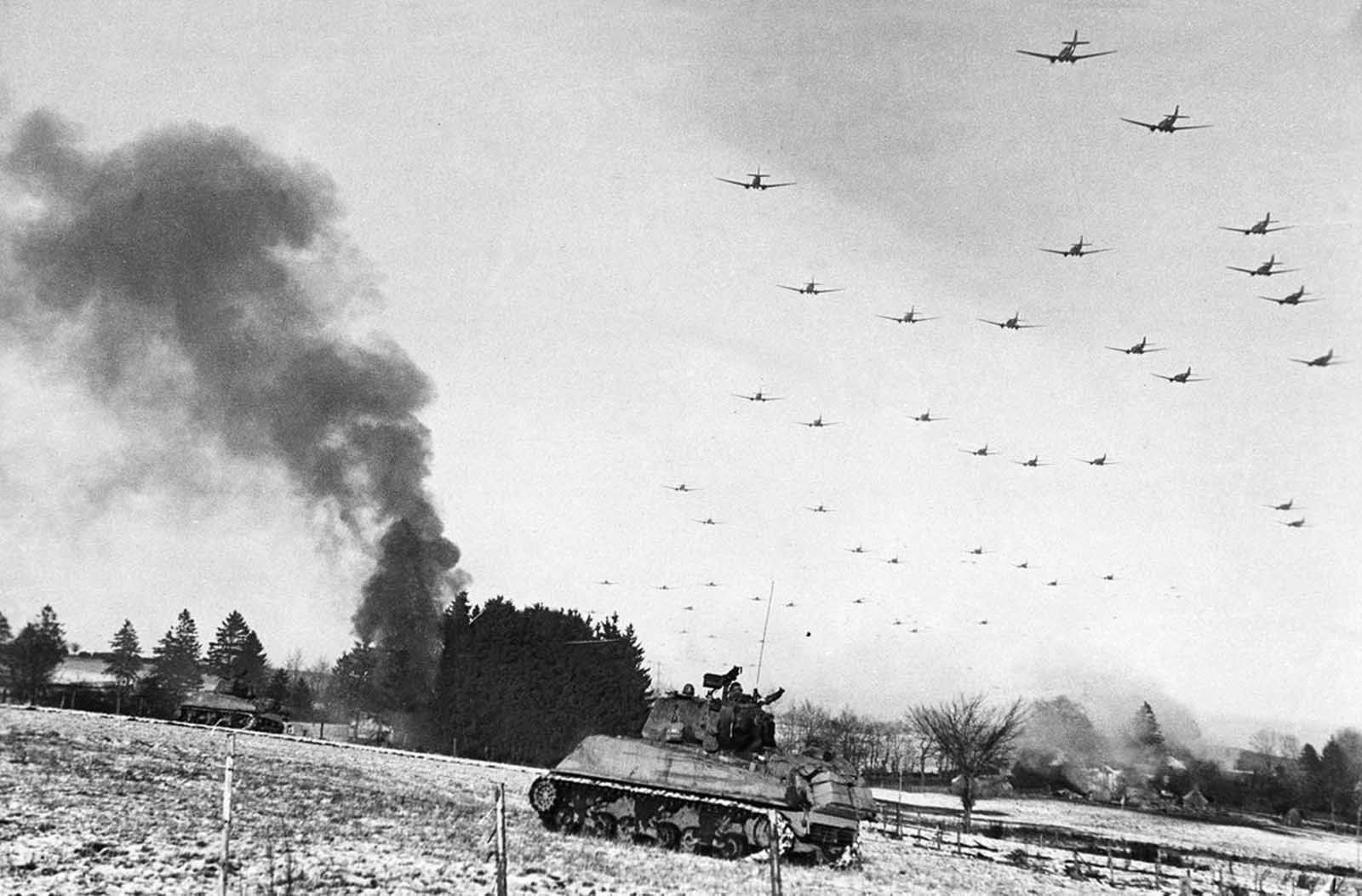 Low flying C-47 transport planes roar overhead as they carry supplies to the besieged American Forces battling the Germans at Bastogne, during the enemy breakthrough on January 6, 1945 in Belgium. In the distance, smoke rises from wrecked German equipment, while in the foreground, American tanks move up to support the infantry in the fighting.