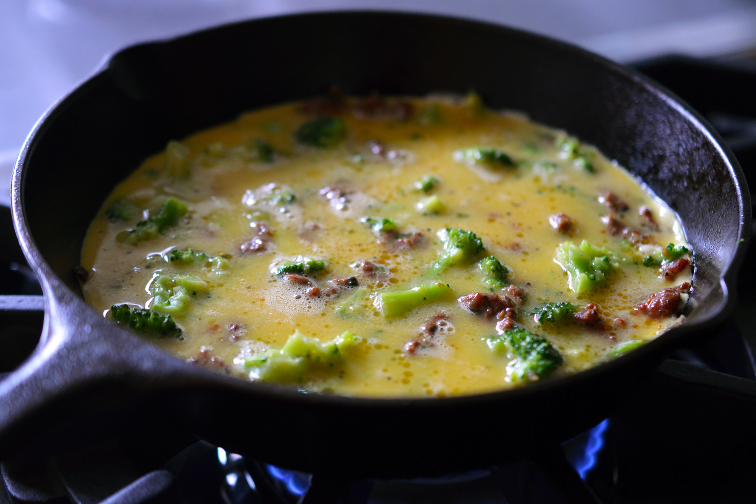 A small cast iron skillet on top of a gas range that is filled with broccoli, ground meat, and uncooked eggs. 