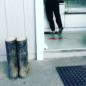 A pair of muddy gumboots outside a fish and chip shop. Inside, the owner of the boots is ordering at the counter.