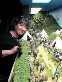 Woman standing next to a diorama of 19th-century soldiers in an encampment in the bush.