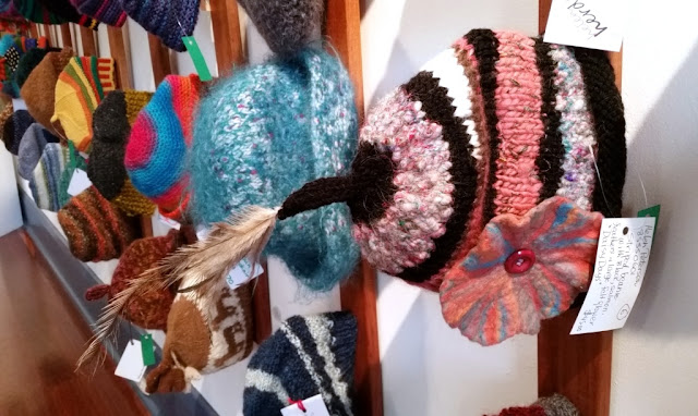 A collection of hats from the western wall of the exhibition. In the foreground is a knitted beanie with a stem on top finished with a natural brown feather. The hat is striped in shades of dark brown, pink and white. The side of the hat is embellished with an appliqued flower which is felted in shades of pink and sky blue and centred with a pink button.