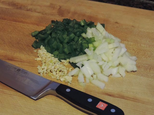 The chopped onion, green pepper, and garlic on the cutting board with a  knife. 