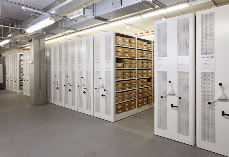 A brightly lit basment room containing ranks of roller Archive shelving.  On an open shelf we can see dozens of brown cardboard boxes all labeled.