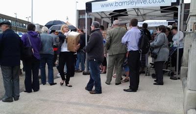 A group of people, mostly facing left stood on a slope, a black and white gazebo labelled Barnsley Live and a woman struggling with a large cardboard box.